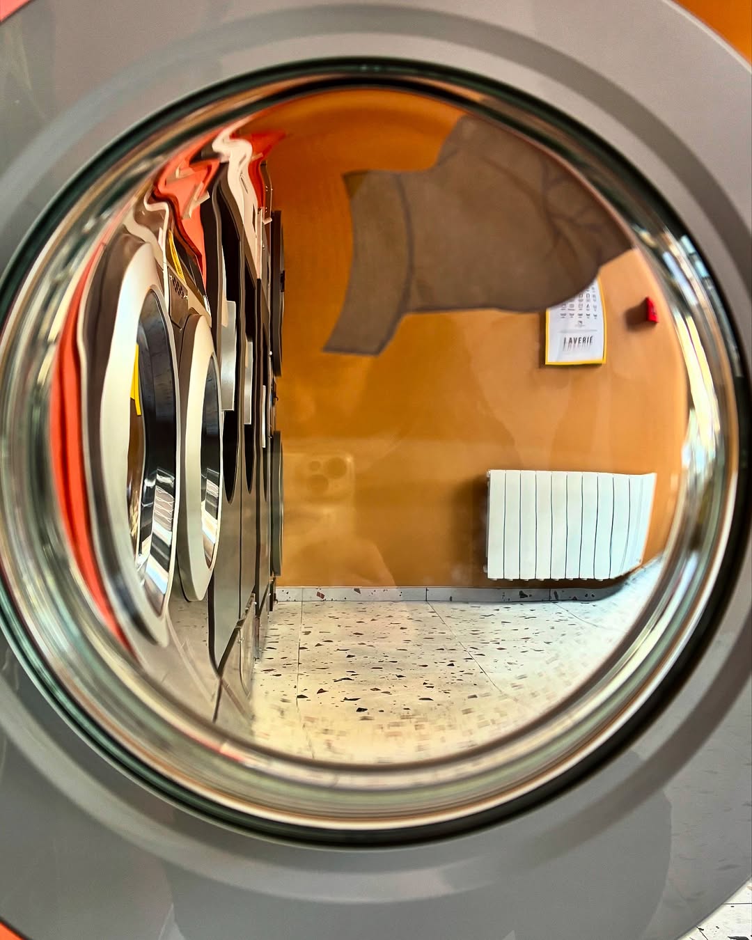 View through a washing machine drum showing the orange-walled laundrette interior at Relax a Wash